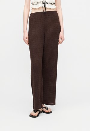 Frau trägt dunkelbraune, hochtaillierte, weit geschnittene, strukturierte Hose mit schwarzen Sandalen und einem weißen, bauchfreien Top vor einem einfarbigen Hintergrund.