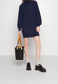 Marinblå stickad polokragsklänning med långa ärmar, tillsammans med svarta kraftiga skor och en svart toteväska med bruna handtag.