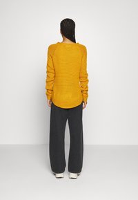 Maglione oversize giallo con un design a maglia testurizzata, polsini a coste e orlo arrotondato, abbinato a pantaloni grigi larghi e sneakers.