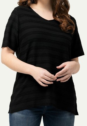 Kvinde iført en sort, tekstureret kortærmet top med vandrette striber, kombineret med blå jeans, hænder løst foldet ved taljen.