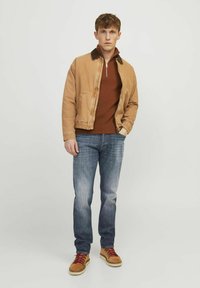 Chaqueta de algodón marrón con un cuello oscuro, combinada con un suéter de cremallera color óxido y jeans de denim azul. Zapatos marrones con cordones rojos.
