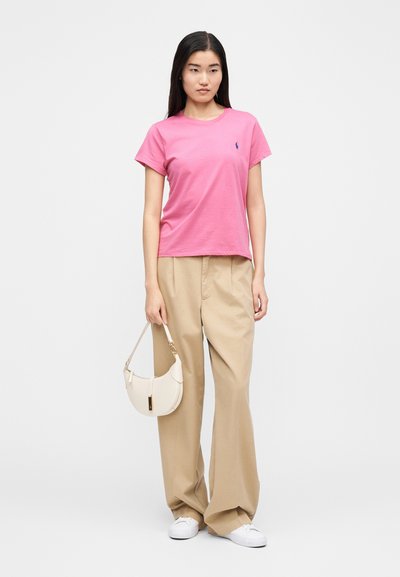 Rózsaszín pamut póló rövid ujjakkal és kerek nyakkivágással, bézs színű, bőszárú nadrággal kombinálva. Kiegészítők között egy kis krémszínű kézitáska található.