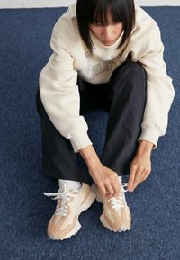 Beige und weiße Sneakers mit einem strukturierten Obermaterial und einer gepolsterten Sohle. Die Schnürsenkel werden gebunden, während man auf einem blauen Teppich sitzt.