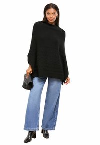 Poncho nero lavorato a maglia con motivo testurizzato, collo alto e spacchi laterali, abbinato a jeans in denim a gamba larga e stivali neri con tacco.