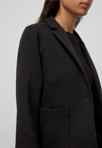 Blazer negro con solapas de pico, cierre de un botón y bolsillos frontales. La textura de la tela parece suave y ligeramente elástica.