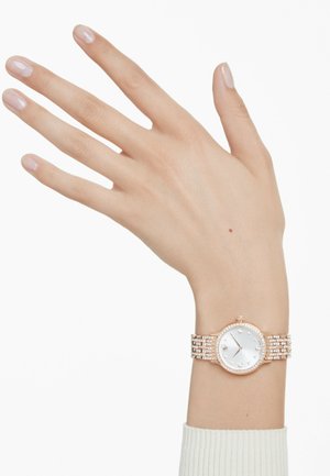 Roze gouden horloge met een ronde kast, witte wijzerplaat en cristallen accenten. De armband heeft een stijlvormen met meerdere schakels en een gestructureerde afwerking.