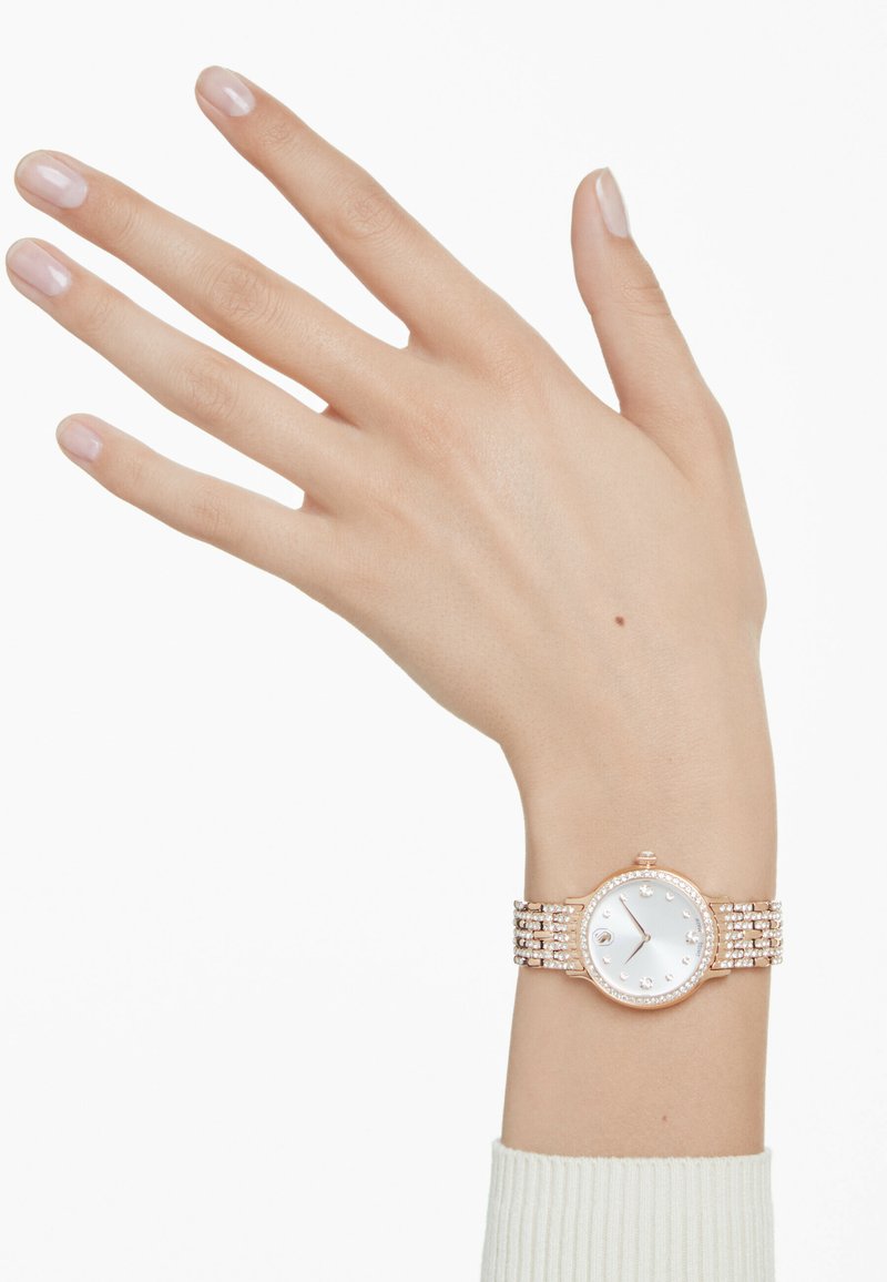 Złoty zegarek różowego koloru z okrągłą kopertą, białą tarczą i kryształowymi akcentami. Bransoletka w stylu ogniwowym z wieloma ogniwami i teksturowanym wykończeniem.