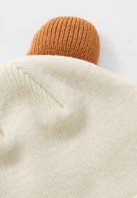 Gestrickte Mütze in Elfenbein mit einer strukturierten Oberfläche, die an der Oberseite ein geripptes, orangefarbenes Detail aufweist.