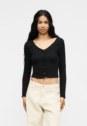 Femme aux longs cheveux foncés portant un haut court noir boutonné à manches longues et un jean blanc taille haute sur un fond uni.