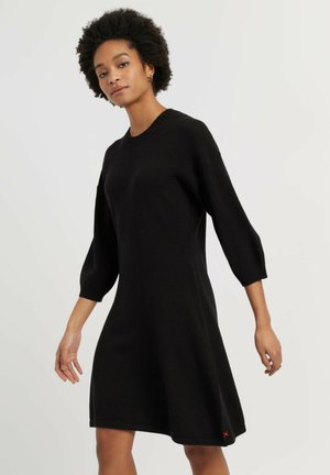 Donna con capelli ricci naturali che indossa un vestito maglione nero al ginocchio con maniche tre quarti, in piedi su uno sfondo semplice.