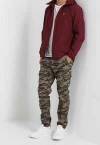 Bordowa lekka kurtka, biała koszulka, spodenki cargo w kamuflażu oraz białe sneakersy. Kurtka ma zamek błyskawiczny i kaptur.