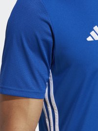Kék sportpóló légáteresztő, textúrált anyagból. Fehér csíkok futnak végig az oldalán, és egy fehér logó található a mellkasán.
