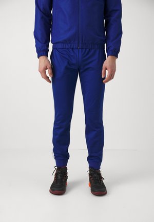 Mann trägt einen blauen Trainingsanzug mit Reißverschluss und elastischen Bündchen sowie schwarze Sneakers mit roten Akzenten, steht vor einem weißen Hintergrund.