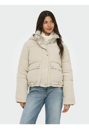 Femme portant une doudoune en velours côtelé beige clair avec des poches et un col montant, associée à un jean bleu et un pull en tricot blanc.