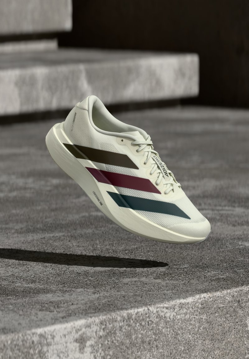 Λευκό αθλητικό παπούτσι με μαύρες, κόκκινες και μπλε ρίγες που αιωρείται πάνω από σκυρόδεμα με τις λέξεις "Adizero" και "Lightstrike Pro" ορατές.