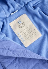 Giacca in pile azzurro chiaro con una consistenza morbida, dotata di un'etichetta bianca interna per i nomi e un design a stella. Bordo accentato in un blu leggermente più scuro.
