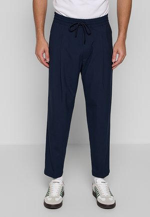 Man draagt marineblauwe broek met trekkoord, witte sokken en witte sneakers met groene strepen, staand tegen een lichtgrijze achtergrond.