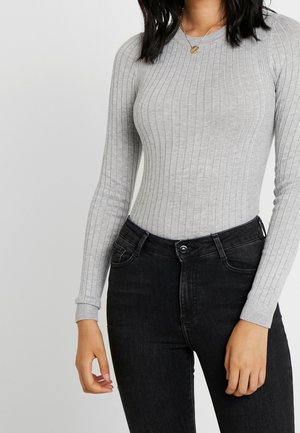 Femme portant un haut gris clair côtelé à manches longues ajusté, rentré dans un jean noir taille haute, accessoirisée d'un collier pendentif en or.