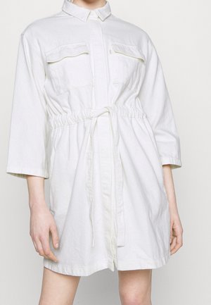 Kvinde iført en hvid skjortekjole med lange ærmer, brystlommer og bindebånd i taljen, vist fra skuldre til midt på låret mod en ensfarvet baggrund.