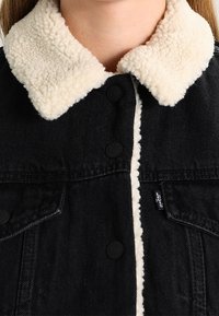 Chaqueta de mezclilla negra con cuello de borrego sintético en color crema, cierre de botones y bolsillos laterales. Cuenta con una etiqueta de marca visible en el lateral.