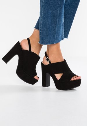 Czarne zamszowe szpilki na platformie z grubym blokowym obcasem, otwartym palcem i krzyżującym się paskiem, z regulowanym paskiem wokół kostki.