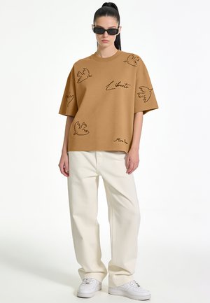 Femme portant un t-shirt marron oversize avec des motifs d’oiseaux et le texte "Liberté", un pantalon large couleur crème, des baskets blanches et des lunettes de soleil noires.