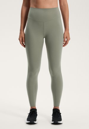 Olijfgroene high-waisted leggings van een gladde, rekbare stof, met een strakke pasvorm en eenvoudig ontwerp. Draagt met zwarte sneakers.