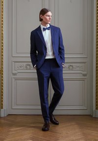 Temno modra suknja z žepnim ovratnikom, bela srajca in usklajena metuljčka; teksturirano blago, oprijet hlače in rjavi usnjeni čevlji.