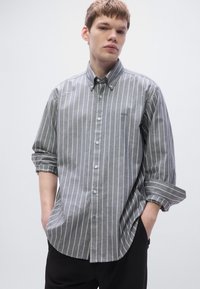 Szürke csíkos, gombos hosszú ujjú ing klasszikus gallérral és felhajtott mandzsettával, fekete nadrágra lazán felhajtva.