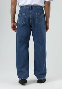 Jean en denim bleu foncé avec une coupe décontractée, deux poches arrière et une coupe droite. Le tissu présente une légère texture. Chaussures noires visibles.