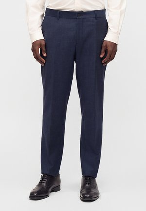 Homme portant un pantalon de costume bleu foncé, des chaussures de ville noires et une chemise blanche à manches longues, se tenant devant un fond clair uni.