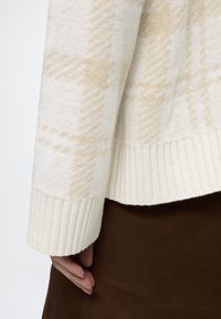 Gros plan d'une main reposant près de l'ourlet d'un pull beige à carreaux avec des poignets côtelés et une jupe marron.