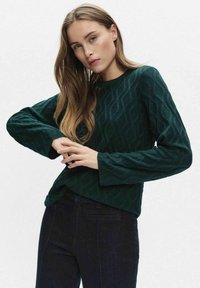 Donkergroene gebreide trui met een textuur van golfpatroon, ronde halslijn en lange mouwen, gecombineerd met een donkere spijkerbroek.