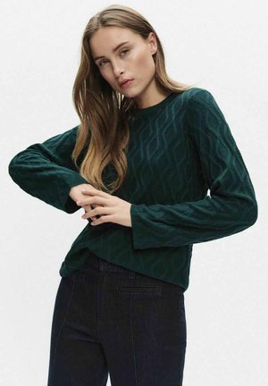 Mörkgrön stickad tröja med ett strukturerat vågmönster, rund halsringning och långa ärmar, kombinerad med mörka jeans.