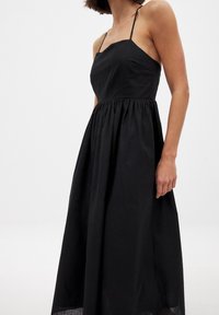 Mustasuora, hihaton maksimekko, jossa on vartalonmyötäinen yläosa ja kerrottu vyötärö. Ominaisuuksiin kuuluvat ohuet olkaimet ja virtaava, teksturoitu hame.