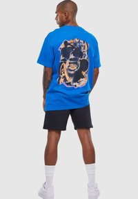 Blå bomuld T-shirt med et stort grafisk print på ryggen, der viser ansigtstræk i sorte og orange nuancer, parret med sorte shorts og hvide sneakers.
