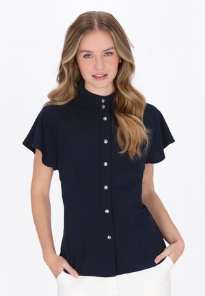 Młoda kobieta z długimi falującymi włosami nosi granatową bluzkę na guziki z krótkimi, falującymi rękawami, stoi z rękami w kieszeniach białych spodni.