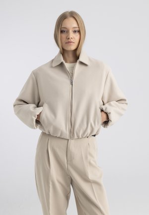 Jeune femme aux cheveux blonds raides portant une veste beige zippée et un pantalon taille haute assorti, debout les mains dans les poches.