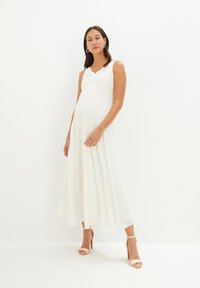 Abito lungo bianco con un corpino in pizzo e una gonna fluente. L'abito presenta una scollatura a V ed è abbinato a sandali nudi con tacco alto.