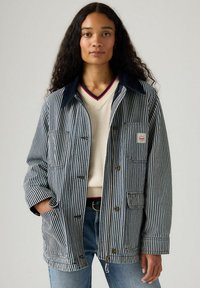 Gestreepte denimjack met een donkerblauwe kraag, voorknopjes en zakken. Draagt over een crèmekleurige trui met een rode accente rondom de halslijn.