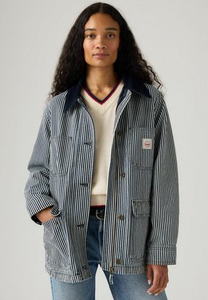 Randig denimjacka med en mörkblå krage, framknappar och fickor. Bärs över en krämfärgad tröja med en röd-accenterad halsringning.