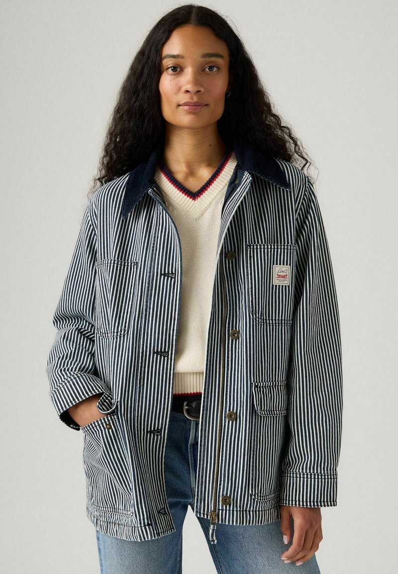 Chaqueta de mezclilla a rayas con un cuello azul oscuro, botones frontales y bolsillos. Llevada sobre un suéter crema con un escote acentuado en rojo.