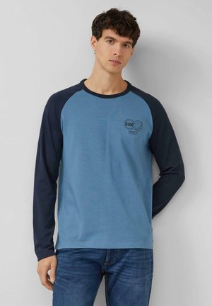 Junger Mann mit lockigem dunklem Haar, der ein blaues und navyfarbenes Langarmshirt mit einem kleinen Emblem auf der Brust trägt und vor einer schlichten weißen Wand steht.