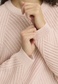 Ljusrosa stickad tröja med ribbat mönster. Har en rund halsringning och ett diagonalt mönster över tyget. Ärmsluten är också ribbade.