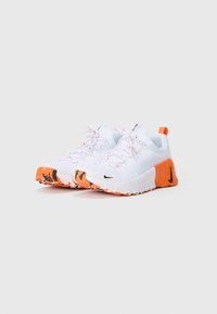 Sapatos de desporto brancos com solas de borracha laranja e pretas, atacadores brancos com manchas laranjas e logótipos da Nike em preto.