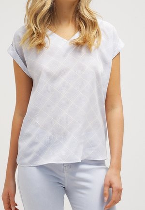 Kvinde iført en hvid kortærmet V-halsbluse med et subtilt ternet mønster og lyseblå jeans.