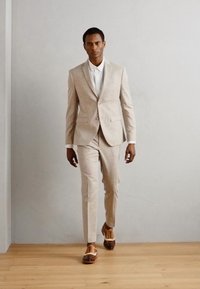 Completo beige su misura con pantaloni slim-fit, camicia bianca e scarpe di pelle marrone con accenti bianchi. Texture liscia e design moderno.