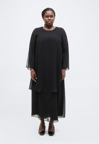 Γυναίκα όρθια μπροστά σε λευκό φόντο, φοράει μακρύ μαύρο φόρεμα με διάφανα μανίκια και μαύρα λουστρίνια παπούτσια με χαμηλό τακούνι.