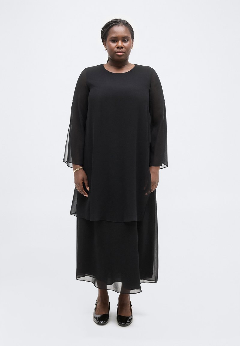 Γυναίκα όρθια μπροστά σε λευκό φόντο, φοράει μακρύ μαύρο φόρεμα με διάφανα μανίκια και μαύρα λουστρίνια παπούτσια με χαμηλό τακούνι.