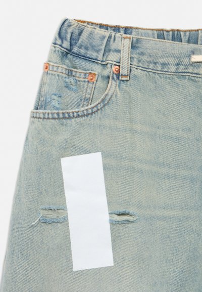 Detailný záber na svetlomodrú rifľovú sukňu v opotrebovanom štýle s predným vreckom, medenými nitmi a bielou obdĺžnikovou štítkom na látke pri horizontálnom trhaní.